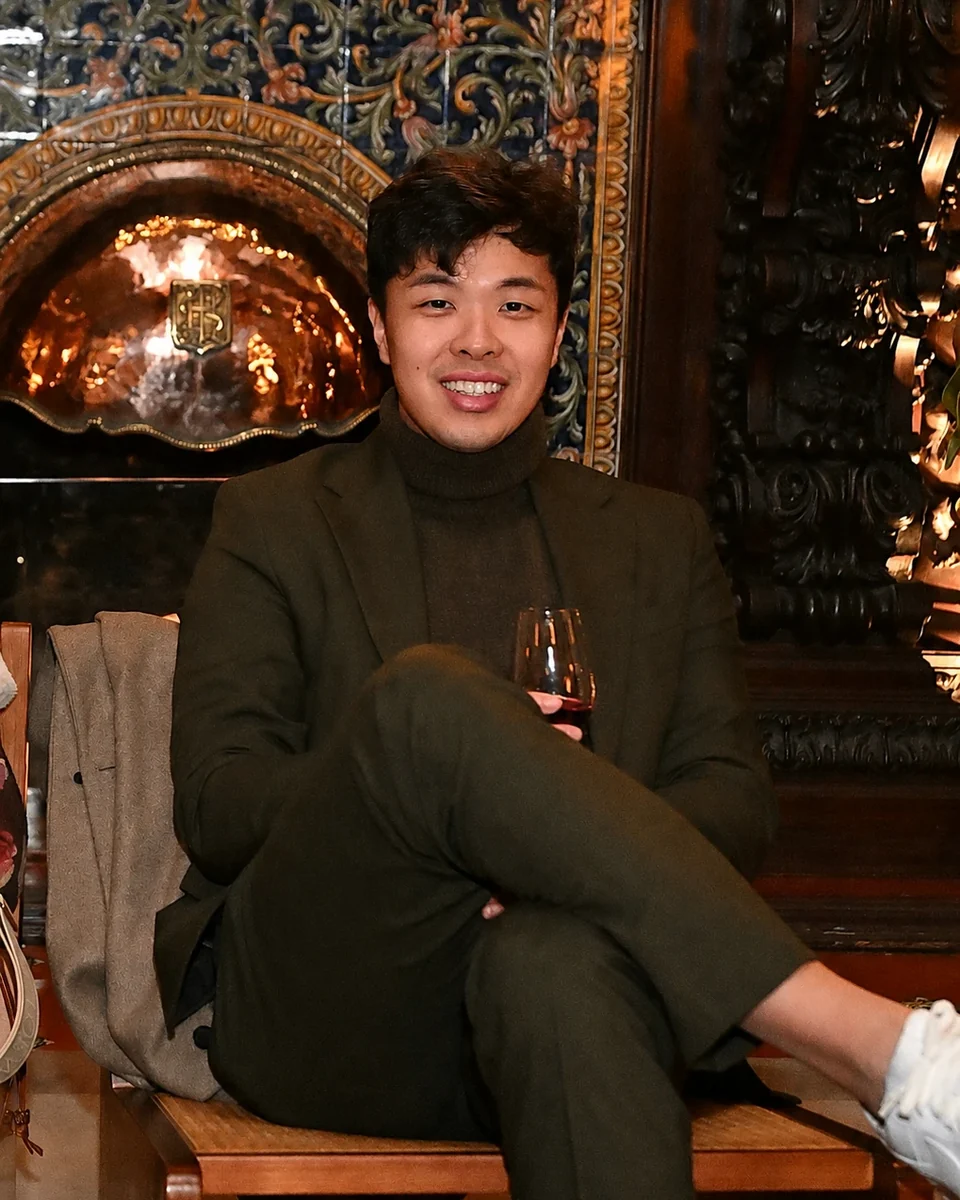 Booker Liao in a dark green suit, seated by a tiled fireplace in Lisbon